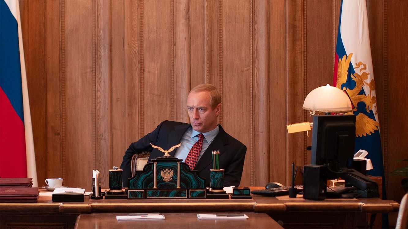 Man in een bureauomgeving, zittend achter een bureau met nationale vlaggen en documenten.