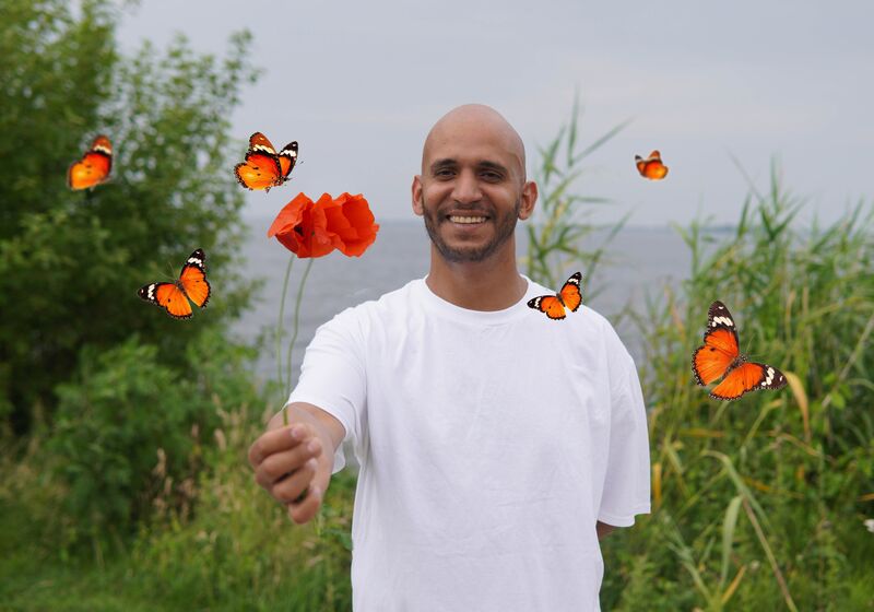 Man in wit T-shirt staat in de natuur en houdt een rode bloem vast, omringd door vlinders.