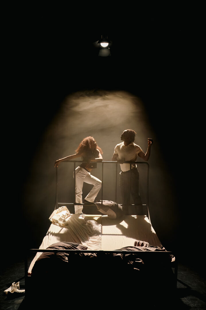 Twee acteurs in theatrale kleding op een bed, verlicht door een enkele spot, met een dramatische sfeer.